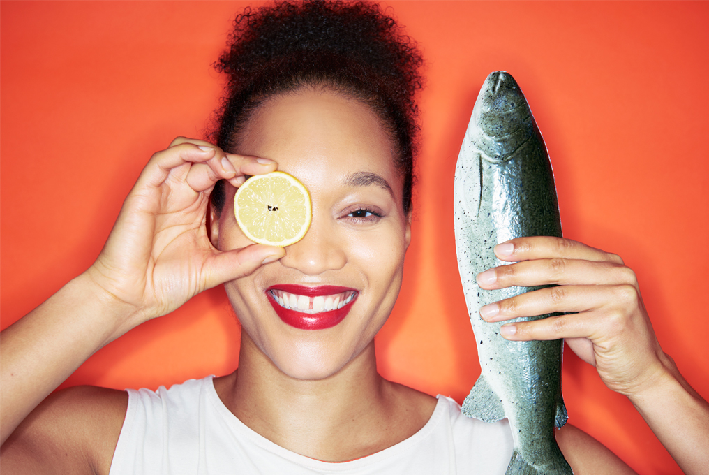 Eine Frau die in der linken Hand einen Fisch und in der rechten Hand vor ihrem Auge eine Zitronenscheibe hält.