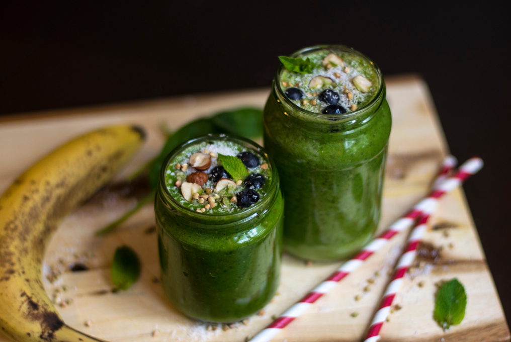Zwei Gläser mit grünem Inhalt auf einem Tablett mit einer Banane Neben dran und auf dem grünen Inhalt sieht man verschiedene Beeren und Samen.