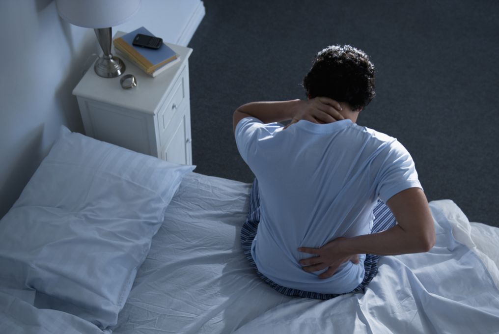 Mann sitzt auf Bett und hält sich am Nacken und Rücken