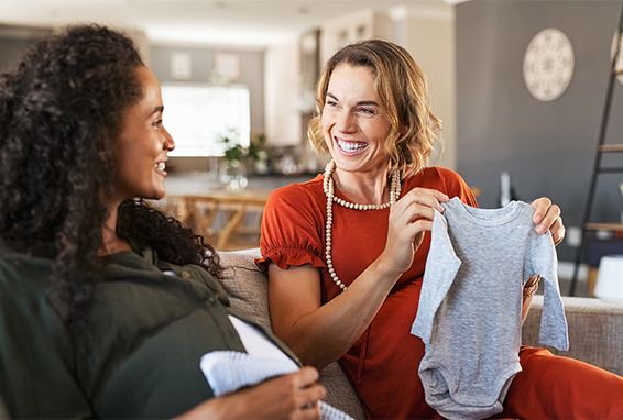 schwangere Frauen auf Sofa mit Babykleidung